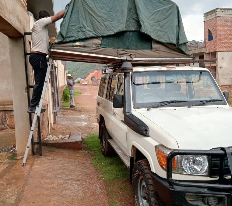 Silver 4x4 utility vehicle with two brown canvas rooftop tents deployed with ladders down on a green grassy lawn surrounded by trees