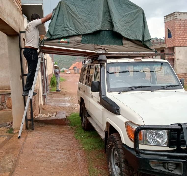 Silver 4x4 utility vehicle with two brown canvas rooftop tents deployed with ladders down on a green grassy lawn surrounded by trees