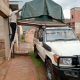 Silver 4x4 utility vehicle with two brown canvas rooftop tents deployed with ladders down on a green grassy lawn surrounded by trees